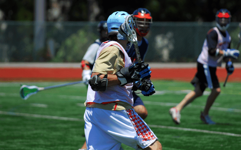 Players practicing lacrosse drills on a field with cones, sticks, and a goal. A coach gives instructions while players run and pass the ball